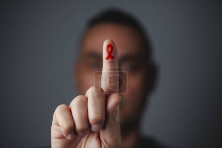 Téléchargez les photos : Un homme a la main devant son visage, avec un ruban rouge de sensibilisation, pour la lutte contre le sida, peint à l'index, sur un fond gris - en image libre de droit