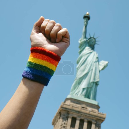 Photo for Closeup of the raised fist of a person, wearing a wristband patterned with the rainbow flag, in front of the Statue of Liberty, New York, United States, in a square format - Royalty Free Image