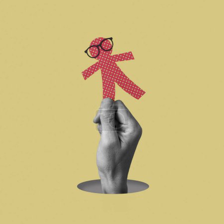 Foto de La mano de un hombre en blanco y negro sostiene una muñeca de papel rojo, con anteojos, sobre un fondo amarillo, como una broma para el día de los inocentes, una fiesta equivalente al día de los tontos de abril - Imagen libre de derechos