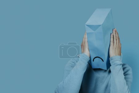 Téléchargez les photos : Gros plan d'un homme avec un sac en papier bleu dans la tête avec une bouche triste dessinée dedans, et ses mains autour, sur un fond bleu avec un peu d'espace vide sur la gauche - en image libre de droit