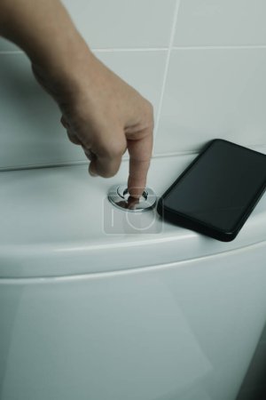Téléchargez les photos : Gros plan d'un homme rincer les toilettes après l'avoir utilisé, et après avoir utilisé son smartphone qui est parti sur le dessus de la citerne - en image libre de droit