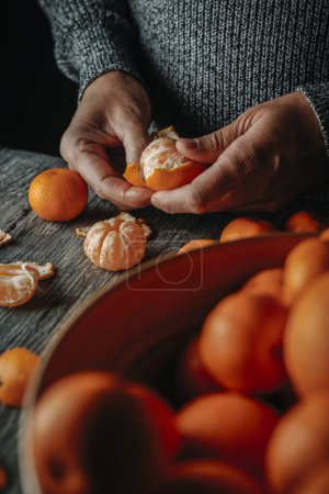 Photo for Closeup of a man, wearing a gray sweater, peeling a mandarin orange on a gray rustic wooden table - Royalty Free Image