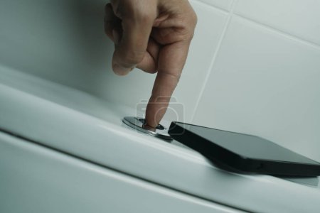 Téléchargez les photos : Gros plan d'un homme appuyant sur le bouton pour rincer les toilettes après l'avoir utilisé et en utilisant son smartphone, à gauche sur le dessus de la citerne - en image libre de droit