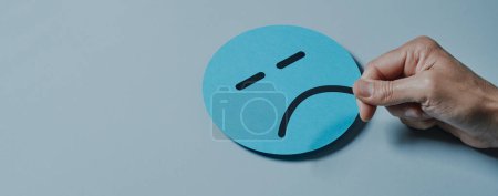 Photo for A man is putting a sad mouth to a blue paper face against a gray background, on a panoramic format to use as web banner or header - Royalty Free Image