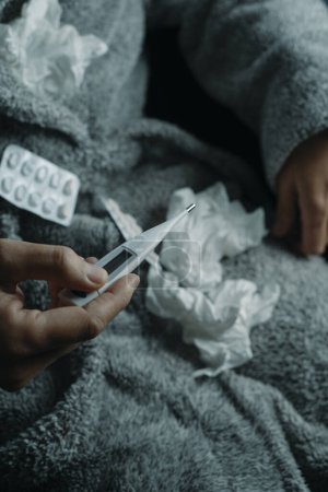 Téléchargez les photos : Un malade à la maison, portant un peignoir gris moelleux, entouré de tissus usagés, est sur le point de mesurer sa température avec un thermomètre - en image libre de droit