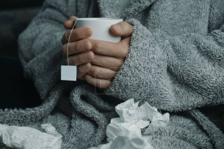 Foto de Un hombre enfermo en casa, con una túnica gris esponjosa, rodeado de pañuelos usados, se calienta con una taza de té caliente de hierbas - Imagen libre de derechos