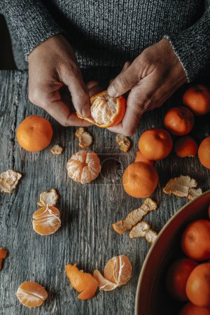 Photo for High angle view of a man, in a gray sweater, peeling a tangerine on a gray rustic wooden table - Royalty Free Image