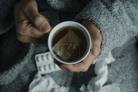 Foto de Detalle de un hombre enfermo, calentándose con una taza de té caliente de hierbas, en casa, usando una túnica gris esponjosa de la casa, - Imagen libre de derechos