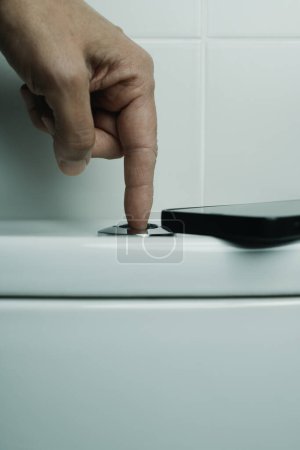 Téléchargez les photos : Un homme appuie sur le bouton sur le dessus de la citerne pour rincer les toilettes après l'avoir utilisé, dans une salle de bain carrelée blanche, avec son smartphone laissé sur le dessus de la citerne - en image libre de droit