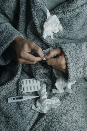 Téléchargez les photos : Un homme caucasien malade à la maison, portant un peignoir gris moelleux, entouré de tissus usagés, est sur le point de prendre une pilule - en image libre de droit