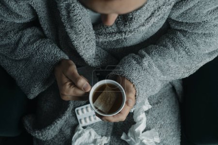 Téléchargez les photos : Vue grand angle d'un homme malade dans une robe de maison grise, à la maison, échauffement avec une tasse de tisane chaude - en image libre de droit