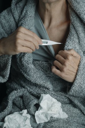 Foto de Un hombre enfermo en casa, vistiendo una túnica gris esponjosa, está a punto de medir su temperatura con un termómetro digital, rodeado de tejidos usados - Imagen libre de derechos