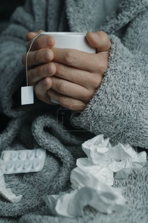 Foto de Primer plano de un hombre enfermo en casa, en una bata de casa gris, rodeado de tejidos usados, calentándose con una taza de té caliente de hierbas - Imagen libre de derechos