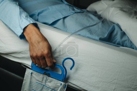 Téléchargez les photos : Gros plan d'un homme, en pyjama bleu, portant un cathétérisme urinaire relié à un sac de drainage couché face au lit - en image libre de droit