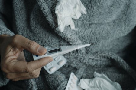 Téléchargez les photos : Gros plan d'un malade à la maison, vêtu d'une robe de chambre gris moelleux, entouré de tissus usagés, sur le point de mesurer sa température avec un thermomètre - en image libre de droit