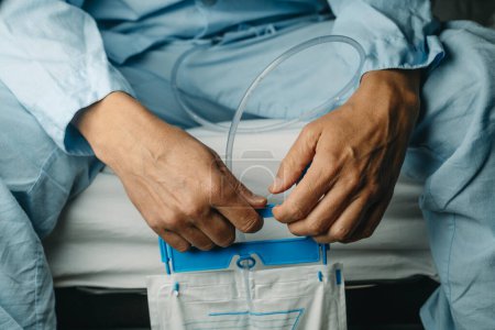 Photo for Closeup of a man, wearing a blue pajamas, holding his urine drainage bag, sitting on the edge of his bed - Royalty Free Image