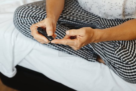 Téléchargez les photos : Un homme, assis les jambes croisées sur le lit, se pique le doigt avec un bâton pour mesurer sa glycémie dans un glucomètre - en image libre de droit