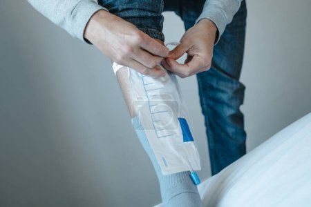 Photo for A young man, wearing casual clothes, attaches a urine drainable bag to his leg using elastic straps - Royalty Free Image