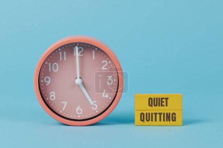 Photo for A pink clock striking five and the text quiet quitting written on two yellow rectangular pieces, on a blue background - Royalty Free Image