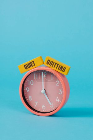 Photo for Closeup of a pink clock striking five and the text quiet quitting written on two yellow rectangular pieces placed on a blue background with some blank space on top - Royalty Free Image