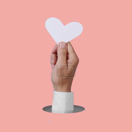 Photo for The hand of a man holding a white heart popping out of a round hole on a pink background - Royalty Free Image