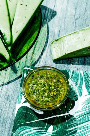 Photo for Closeup of a glass bowl with some pesto sauce next to some slices of pesto cheese in a green glass plate placed on a gray rustic wooden table - Royalty Free Image