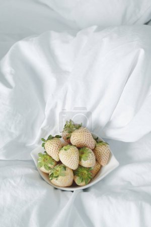 Photo for Some white strawberries in a white ceramic bowl, placed on an unmade bed with daylight - Royalty Free Image