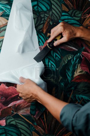 a caucasian man, wearing a gray working coat, is using a paint scraper to remove a strip of floral patterned wallpaper from a wall