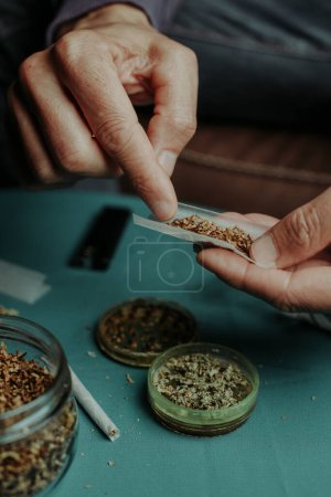Nahaufnahme eines Mannes, der Tabak auf ein Rollpapier legt, um etwas Cannabis hinzuzufügen, sitzt an einem Tisch, an dem eine Mühle mit zerfetztem Cannabis und ein Glas mit rollendem Tabak stehen.