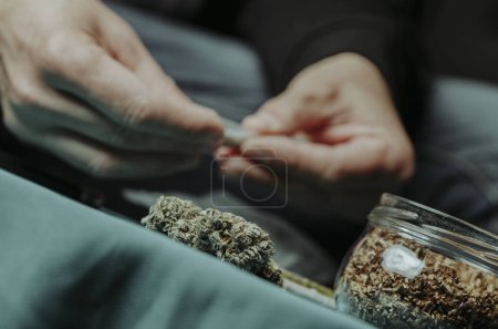 Nahaufnahme eines Mannes, der einen Joint rollt und an einem Tisch sitzt, an dem sich Cannabisknospen und ein Glas mit etwas rollendem Tabak befinden