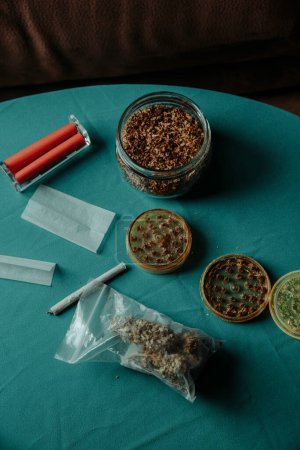 some cannabis buds in a plastic bag on a table next to a used herb grinder, a spliff or joint, some rolling paper sheets and a jar with some rolling tobacco