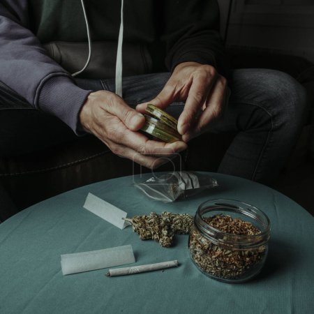 Nahaufnahme eines Mannes, der gerade dabei ist, eine Cannabis-Knospe mit einem gebrauchten Schleifer zu zerkleinern, auf einem Sofa sitzend, neben einem Tisch mit etwas rollendem Tabak und Papier
