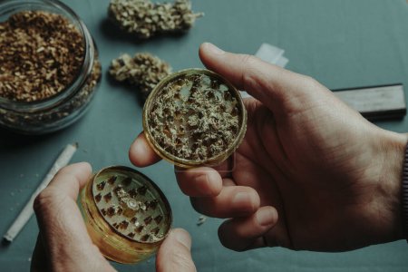 Foto de A man has shredded a cannabis bud with a used grinder, at a table where there is a jar with some rolling tobacco and some more cannabis buds - Imagen libre de derechos