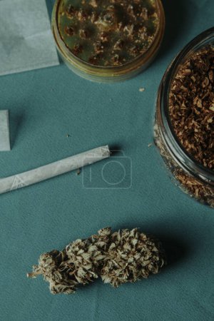 Nahaufnahme einer Cannabis-Knospe auf einem Tisch neben einem gebrauchten Kräutermühle, einem Glas mit etwas Rolltabak und einem Spleiß