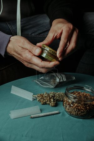 Nahaufnahme eines Mannes, der mit einer gebrauchten Mühle eine Cannabis-Knospe zerkleinert, auf einem Sofa sitzend, neben einem Tisch mit etwas rollendem Tabak und Papier