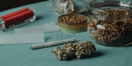 Nahaufnahme einer Cannabis-Knospe auf einem Tisch neben einer gebrauchten Kräutermühle und einem Glas mit etwas rollendem Tabak, im Panoramaformat zur Verwendung als Web-Banner oder Header
