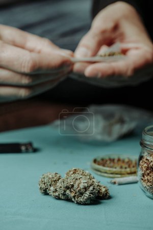 ein Mann rollt einen Joint an einem Tisch, an dem sich einige Cannabisknospen, ein Kräutermahlwerk und ein Glas mit etwas rollendem Tabak befinden
