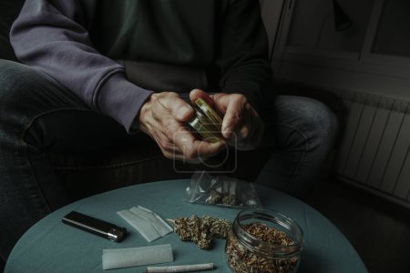 ein Mann, der auf einem Sofa sitzt und eine Cannabisknospe mit einer gebrauchten Mühle zerkleinert, neben einem Tisch mit rollendem Tabak und Papier