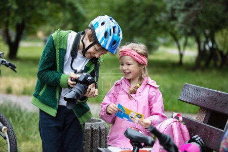 Photo for Young boy and girl looking at camera display and smiling. Brother showing picture he has taking to his younger sister. Children talking and laughing during their bicycle ride in park. - Royalty Free Image