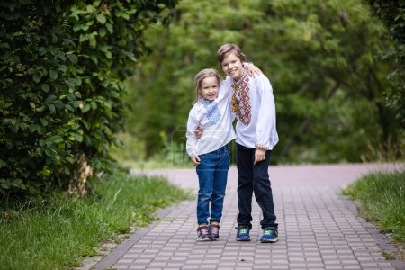 Photo for Young children wearing traditional Ukrainian embroidered shirts hugging each other while looking at camera. Brother and sister posing outdoors in park - Royalty Free Image