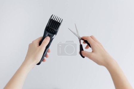 Close up of female hands holding electric hair clipper and scissors on white background