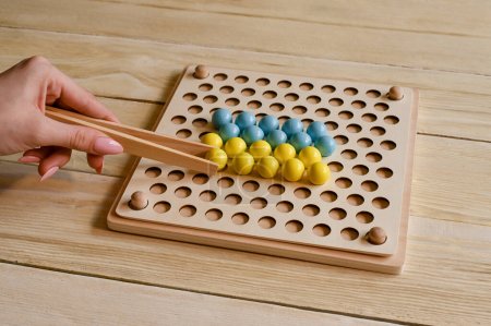 Téléchargez les photos : Wooden Montessori sorting board. In the hands of a wooden tweezers with a ball . The symbol of Ukraine. Develop fine motor skills, logical thinking. - en image libre de droit