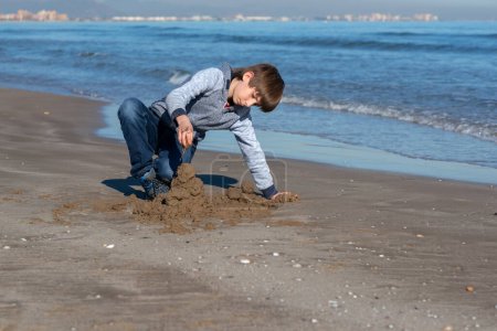 Téléchargez les photos : Jeune beau garçon jouant à la plage d'hiver. Joyeux garçon de 11 ans construisant des châteaux de sable au bord de la mer d'automne. Portrait extérieur du gamin. - en image libre de droit