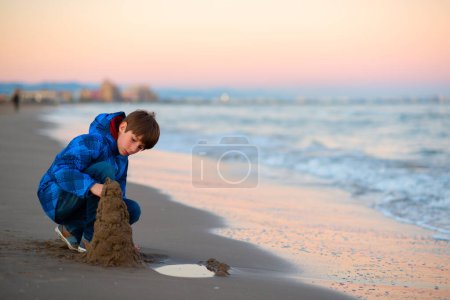 Photo for Young handsome boy playing at the winter beach. Cute happy 11 years old boy building sand castles at seaside. Kid's outdoor portrait. - Royalty Free Image