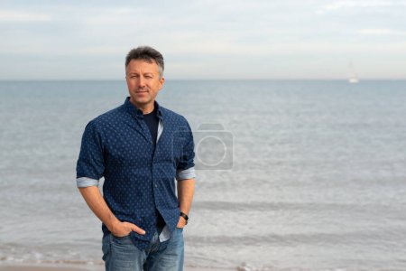 Foto de Al aire libre retrato guapo hombre maduro caminando en la playa. Atractivo modelo masculino adulto posando en la playa en vaqueros azules y camisa. - Imagen libre de derechos