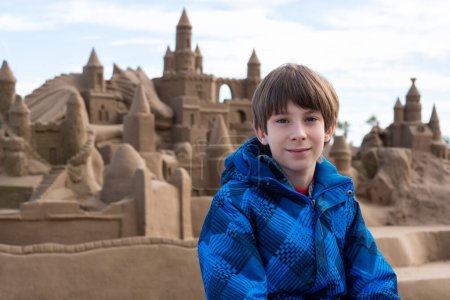 Photo for Young handsome boy posing at the beach over view with sand castle. Cute happy smiling 11 years old boy at autumn seaside. Kid's outdoor portrait. - Royalty Free Image