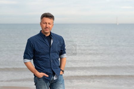 Foto de Al aire libre retrato guapo hombre maduro caminando en la playa. Atractivo modelo masculino adulto posando en la playa en vaqueros azules y camisa. - Imagen libre de derechos