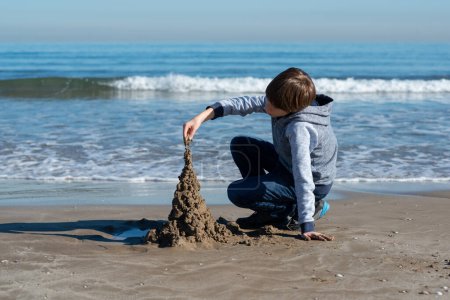 Photo for Young handsome boy playing at the winter beach. Cute happy 11 years old boy building sand castles at autumn seaside. Kid's outdoor portrait. - Royalty Free Image