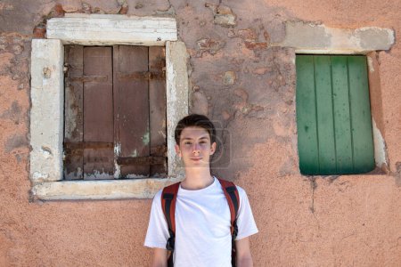 Téléchargez les photos : Beau garçon adolescent posant en plein air sur un vieux mur obsolète avec des fissures dans le plâtre et des fenêtres à volets d'anciens murs à Venise - en image libre de droit