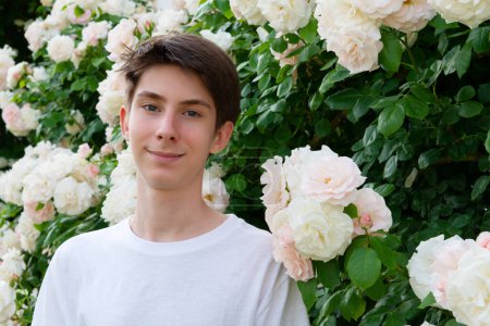 Photo for Teen boy in park among beautiful white-pink blooming roses, summer outdoor - Royalty Free Image